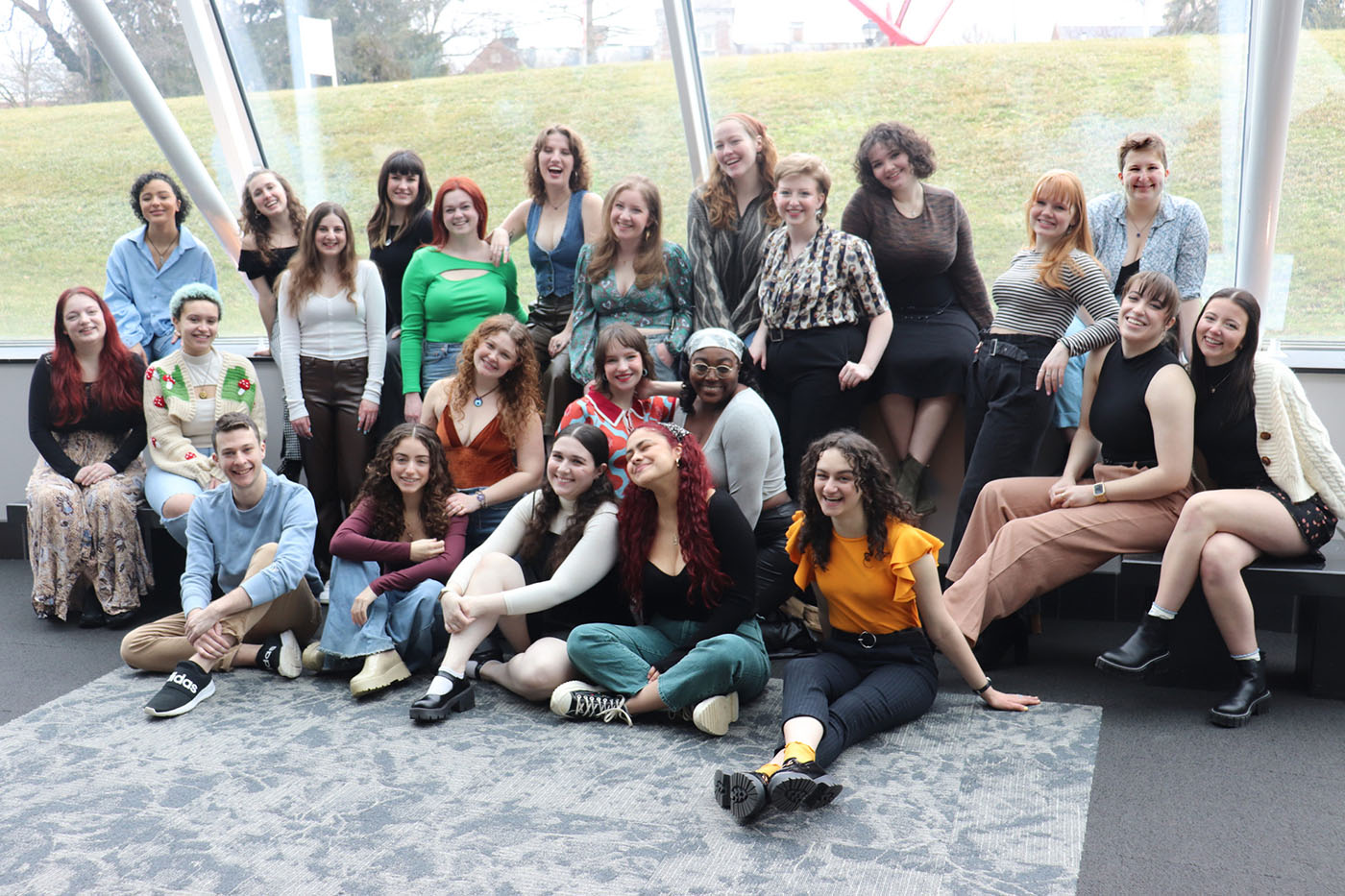 A group of more than 20 college students poses in front of a large window looking out at Muhlenberg's campus
